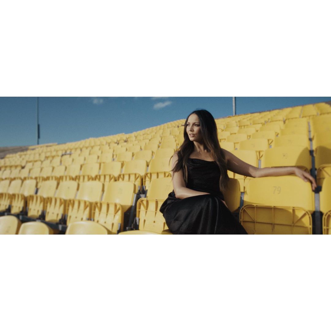 A woman in a black dress sits in the yellow stadium seating on a sunny day under a clear blue sky, looking off to the side.