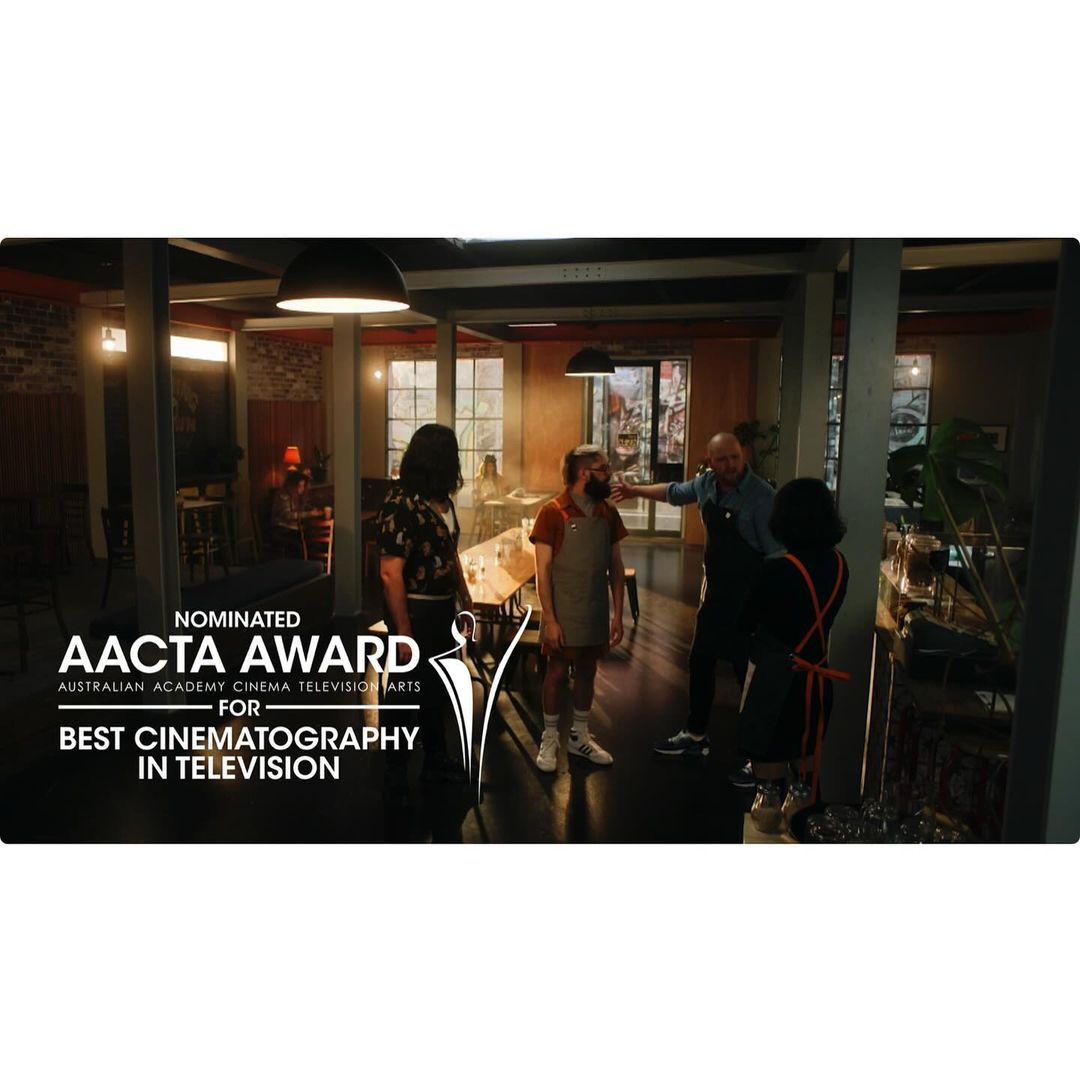 People gathered in a restaurant with an award nomination poster; brick walls, tables, and chairs visible in soft, indoor lighting.