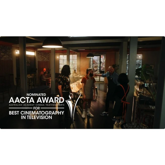 People gathered in a restaurant with an award nomination poster; brick walls, tables, and chairs visible in soft, indoor lighting.