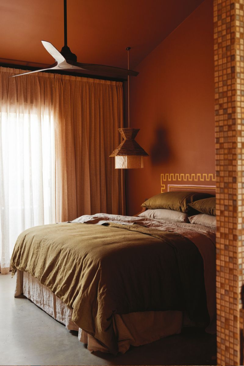 Bedroom interior with a bed covered in a green duvet, a ceiling fan, and an intricate light fixture in an earthy color palette.