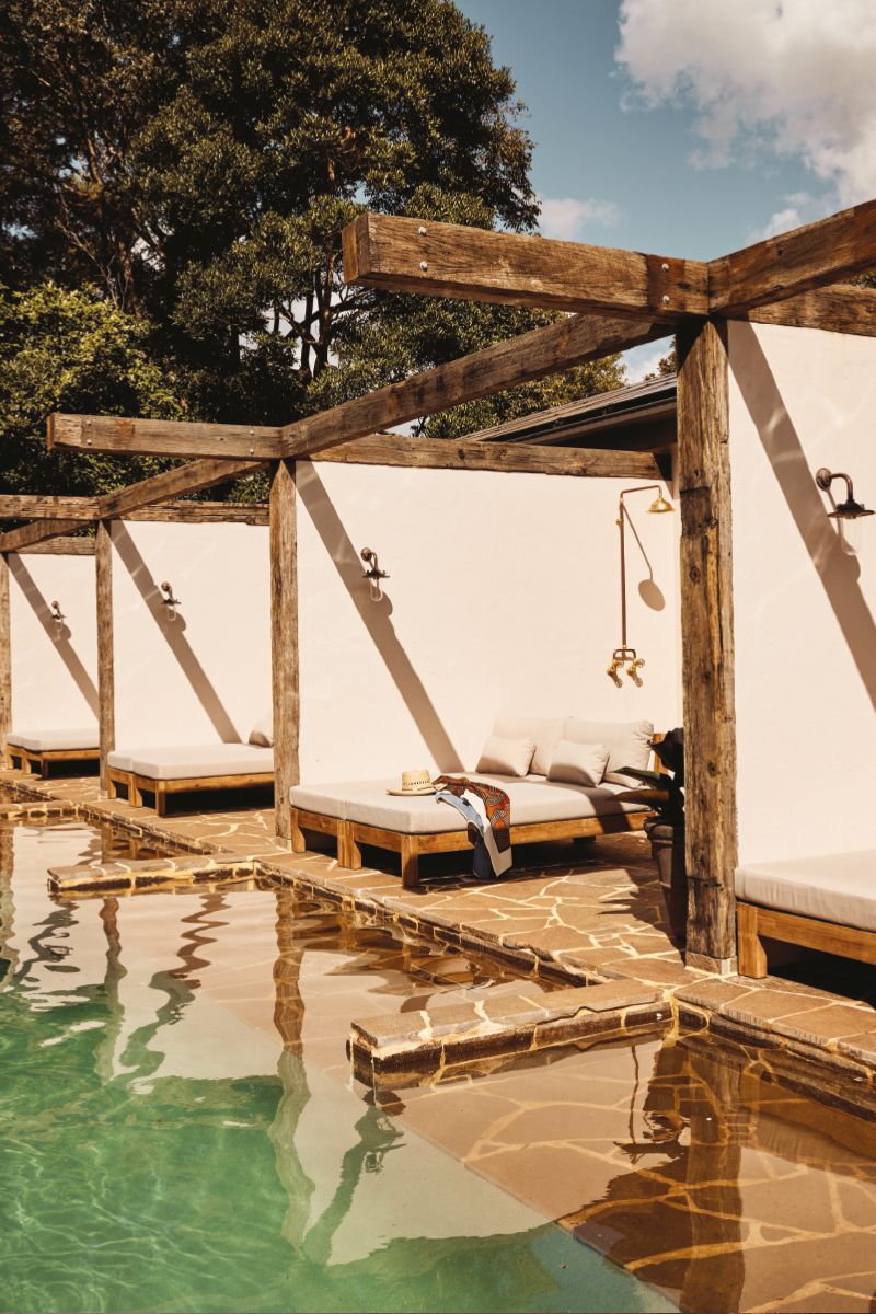 Outdoor pool area with lounge beds and sunbeds under a wooden pergola structure, reflecting on the pool water with blue sky and trees.