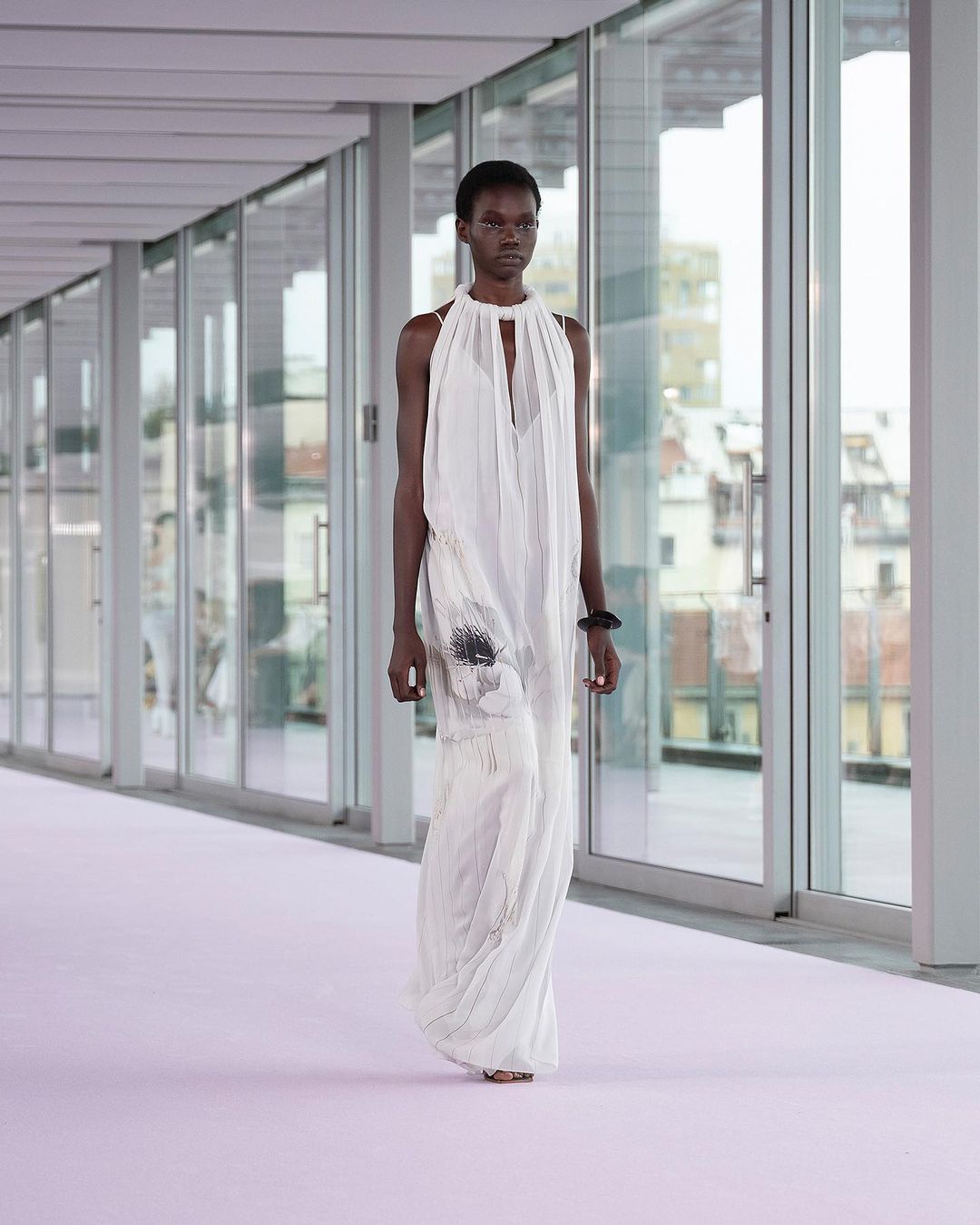 Runway fashion show featuring a black female model in a flowing white halter dress, walking on a light purple runway in an indoor setting.