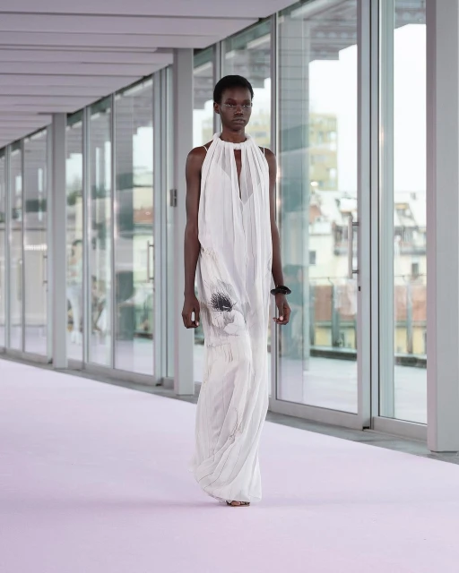 Runway fashion show featuring a black female model in a flowing white halter dress, walking on a light purple runway in an indoor setting.
