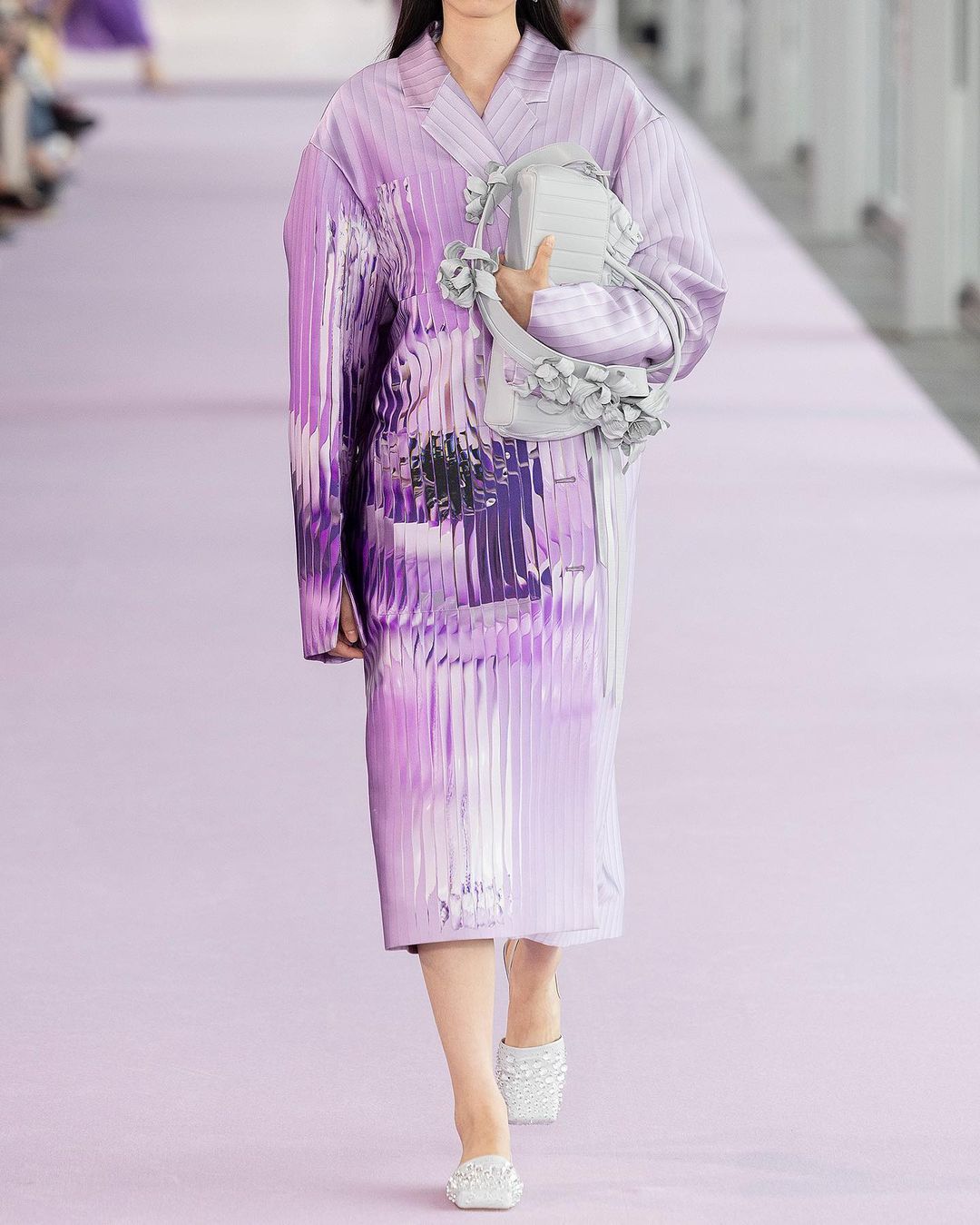 Runway fashion show featuring a model in a purple outfit with a matching purse, standing on a purple runway with spectators in the background.