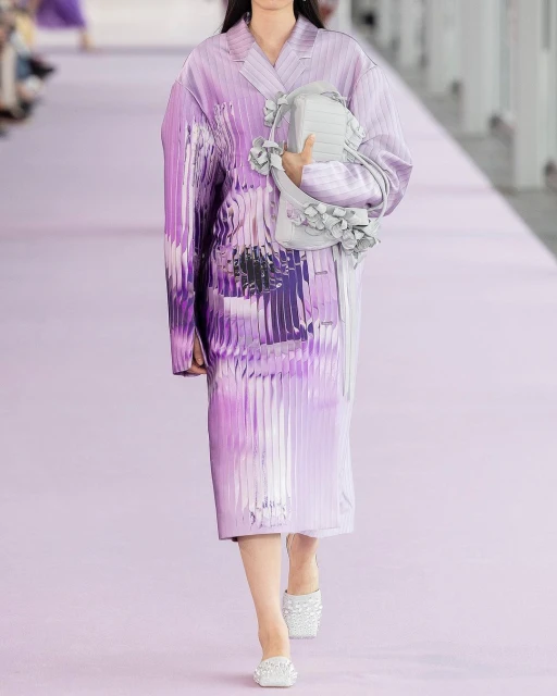Runway fashion show featuring a model in a purple outfit with a matching purse, standing on a purple runway with spectators in the background.