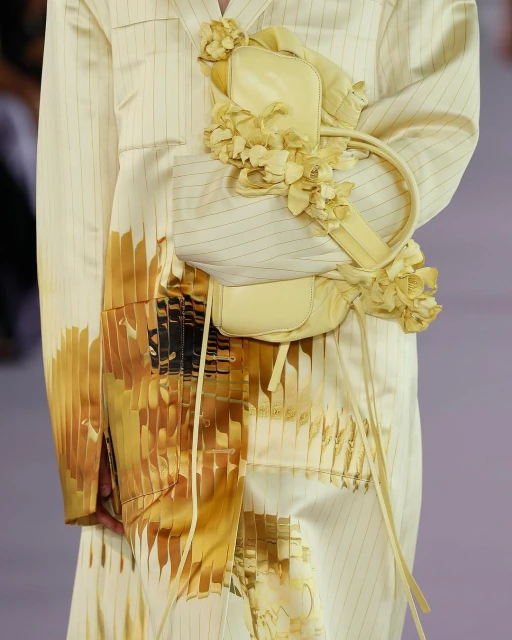 Runway shot of a pale yellow pinstripe jacket with an attached matching bag adorned with flower details, part of a fashion collection.