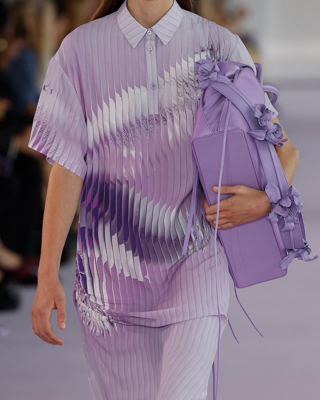 Runway shot featuring a fashion model showcasing a light purple pleated outfit and matching floral-decorated handbag at a fashion show.