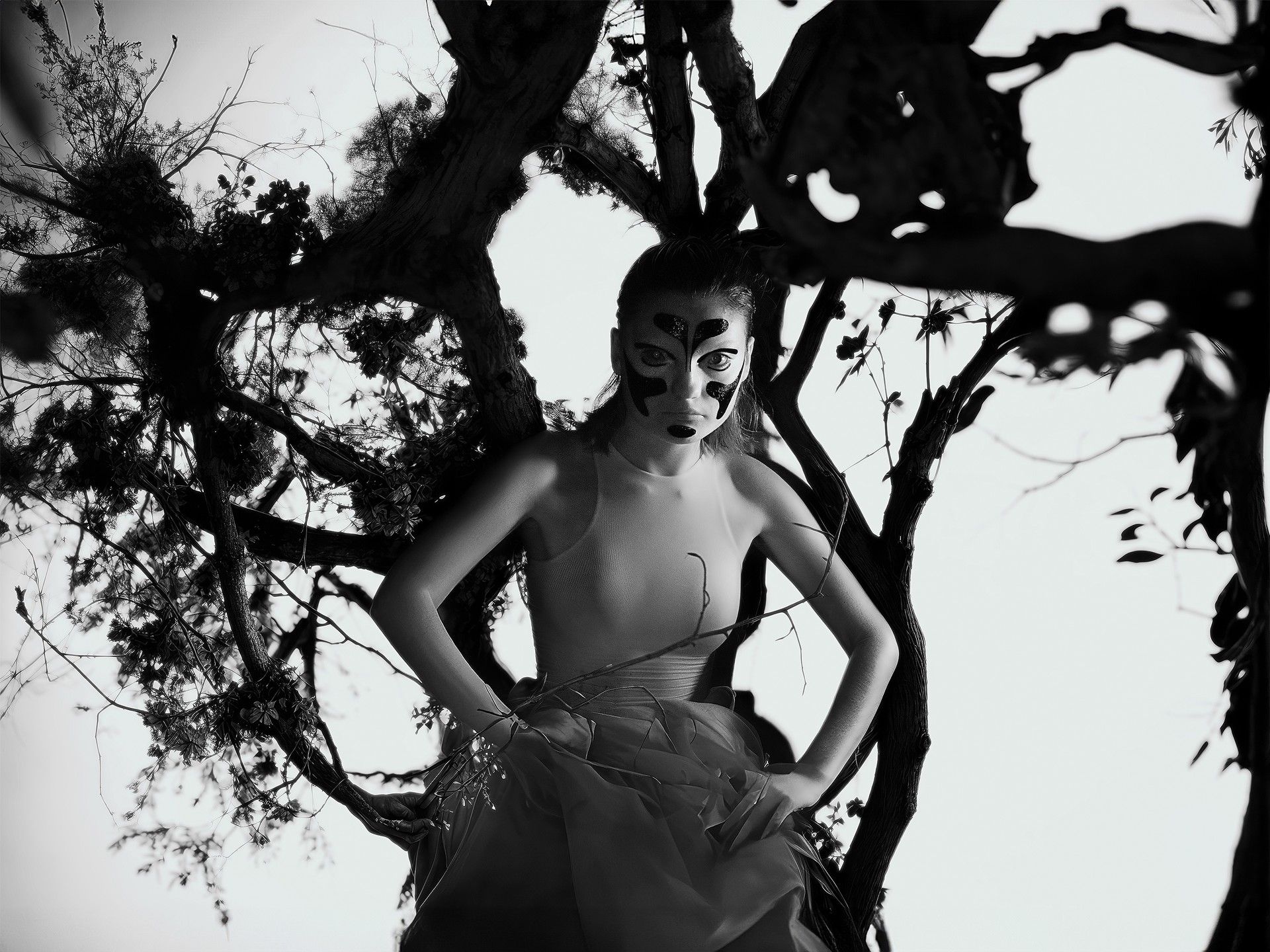 Monochrome portrait of a woman in a light-colored dress and abstract facial makeup posing amidst a dark tree branch structure.