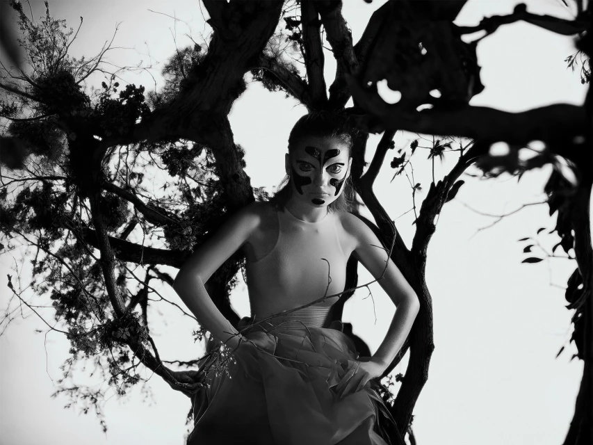Monochrome portrait of a woman in a light-colored dress and abstract facial makeup posing amidst a dark tree branch structure.
