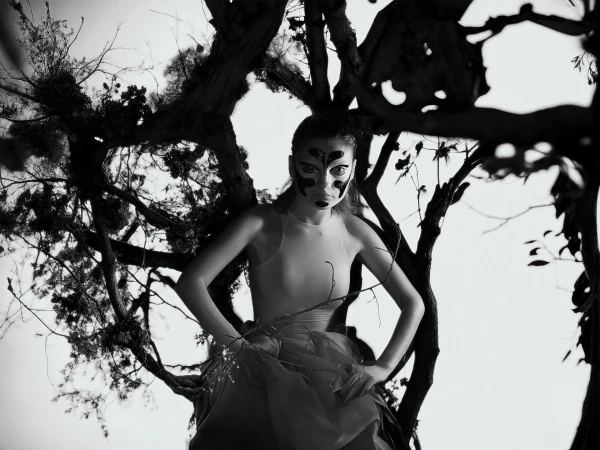 Monochrome portrait of a woman in a light-colored dress and abstract facial makeup posing amidst a dark tree branch structure.