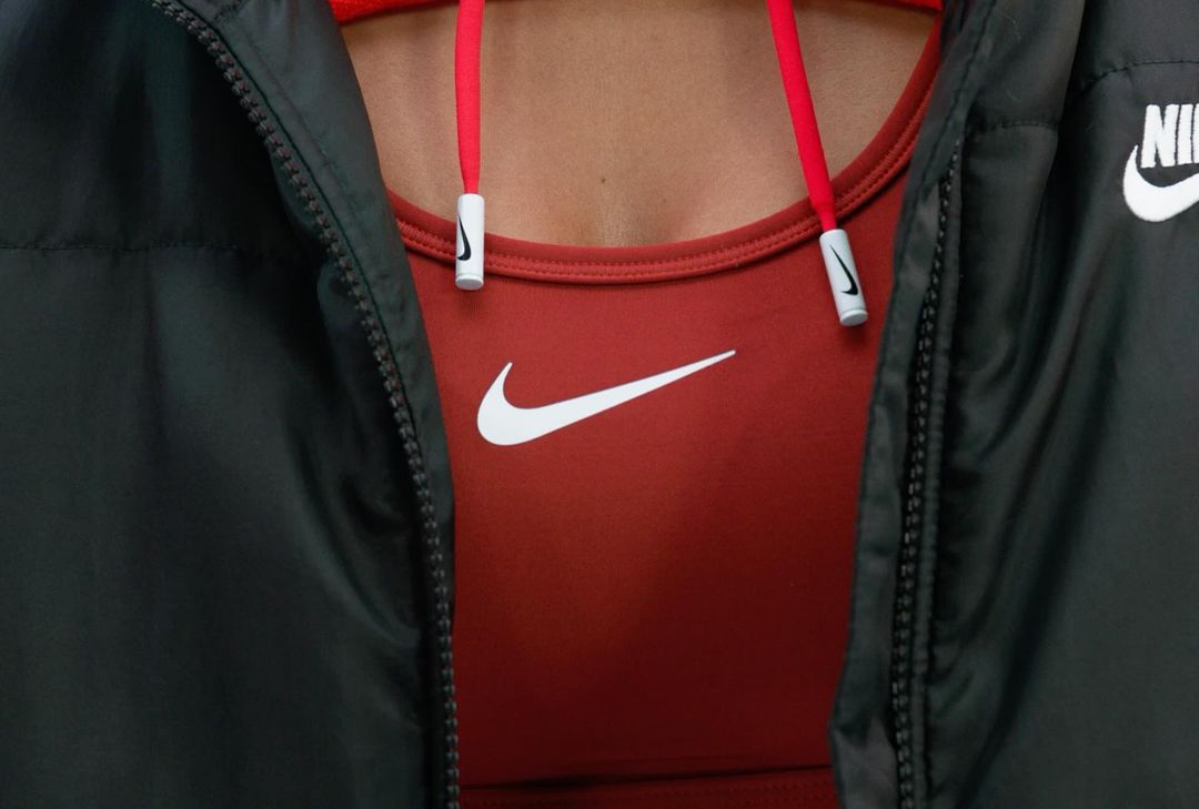 Close-up of a person wearing a rust-colored Nike sports bra with a white swoosh logo, paired with a black Nike jacket, showing the logo.