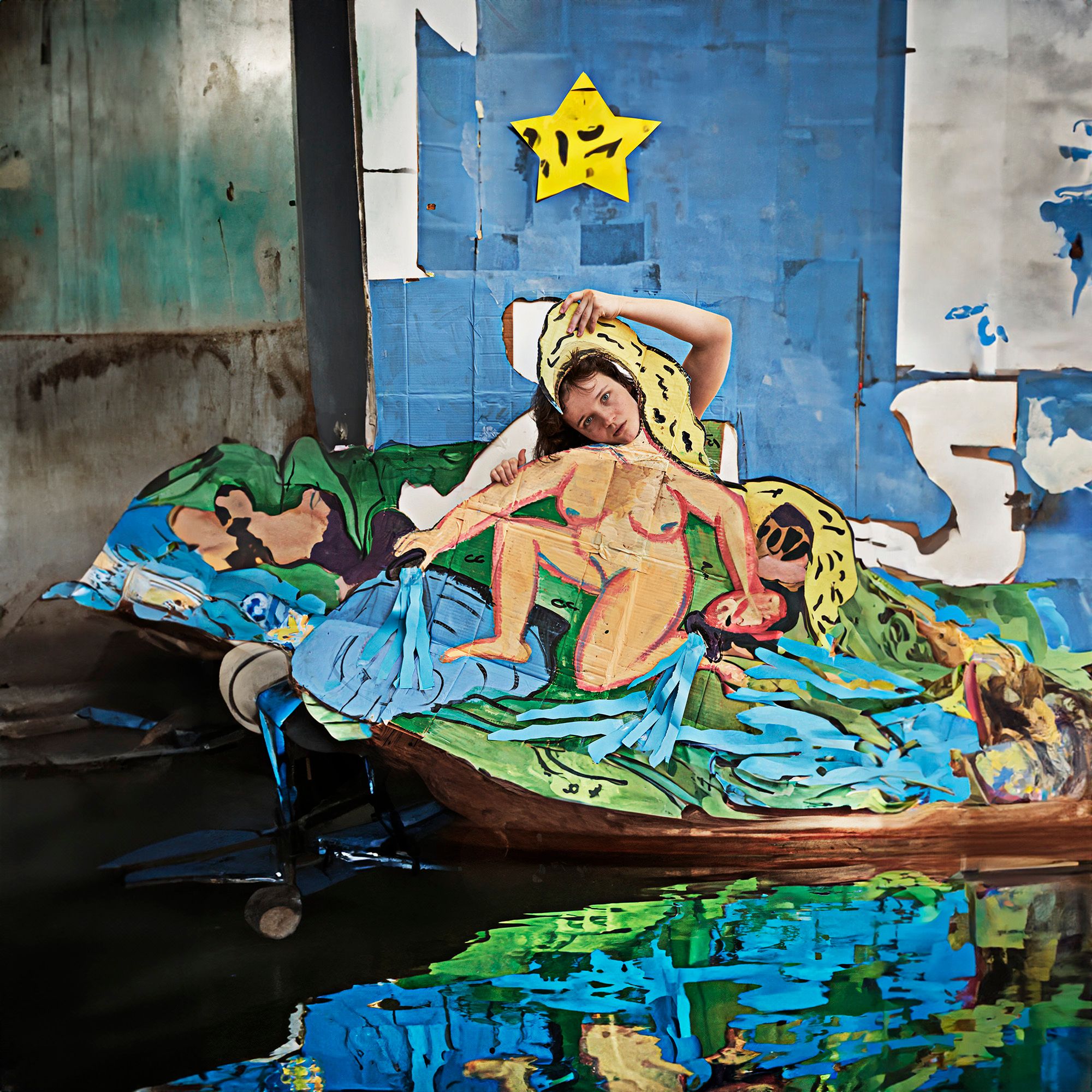 Artistic studio portrait of a red-haired woman in a painted boat on water, wearing a yellow printed shawl, with bright colors.