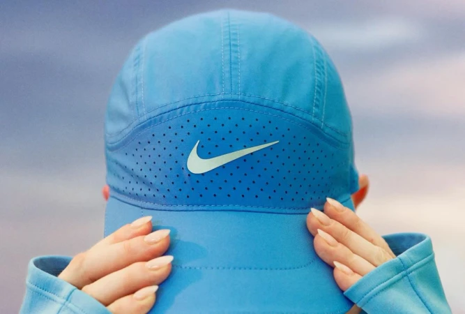 Close-up of a blue baseball cap with a prominent swoosh logo, held by two hands against a soft overcast sky.