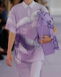 Model wearing a pleated lavender patterned polo shirt carrying a matching lilac handbag with floral appliqués.