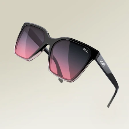 Close-up of black framed sunglasses with a pink and black gradient lens, displaying the brand name QUAY on the side against a beige background.
