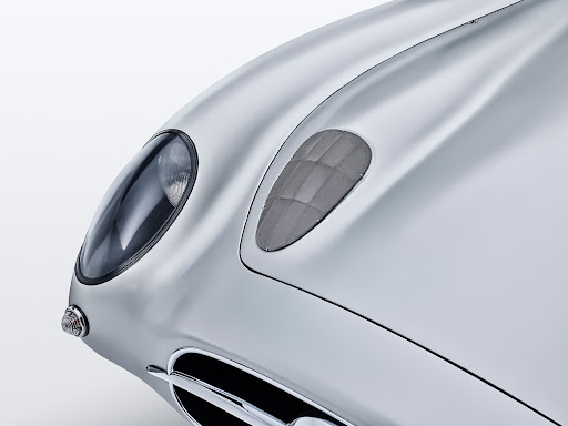 Close-up of a silver vintage car's front with a headlight, vent, and chrome grill against a bright white backdrop in a studio shot.