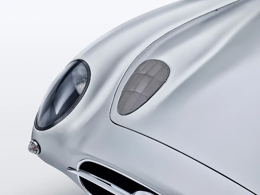 Close-up of a silver vintage car's front with a headlight, vent, and chrome grill against a bright white backdrop in a studio shot.