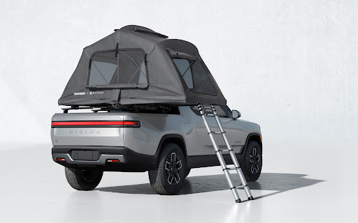 Silver Rivian pickup truck with rooftop tent and ladder, shown in a studio setting. Vehicle is clean and well-lit with a minimalist background.