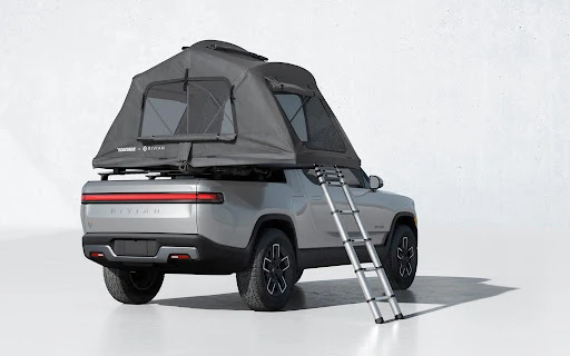Silver Rivian pickup truck with rooftop tent and ladder, shown in a studio setting. Vehicle is clean and well-lit with a minimalist background.