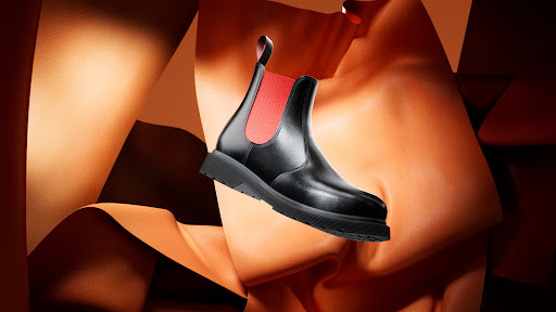 Black leather Chelsea boot with a red accent, set against swirling textures of orange and brown leather in a studio setting.