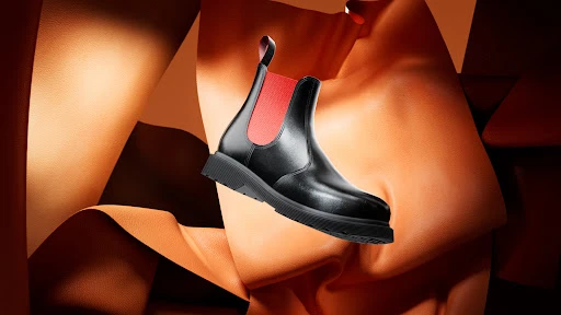 Black leather Chelsea boot with a red accent, set against swirling textures of orange and brown leather in a studio setting.