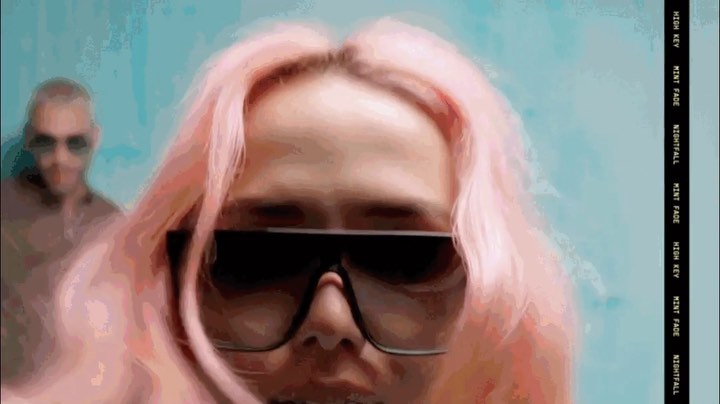 Close-up of a person with pink hair wearing sunglasses, with another person blurred in the background against a light blue wall.