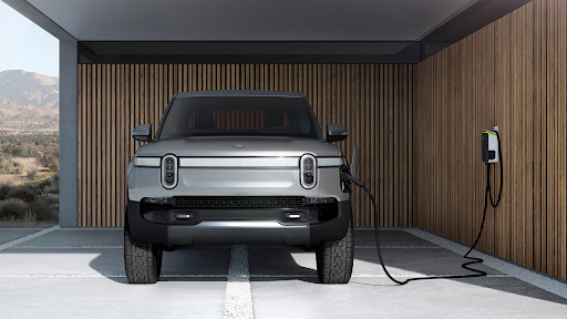 Silver Rivian electric truck charges indoors against wood-paneled wall. Modern home architecture; mountain view through open doorway.
