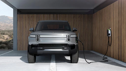 Silver Rivian electric truck charges indoors against wood-paneled wall. Modern home architecture; mountain view through open doorway.