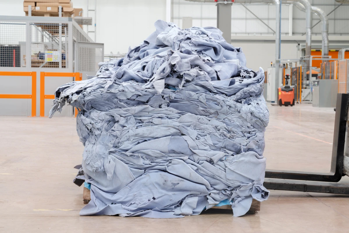 Large pile of folded light blue material on a wooden pallet in a factory or warehouse setting with a forklift nearby.