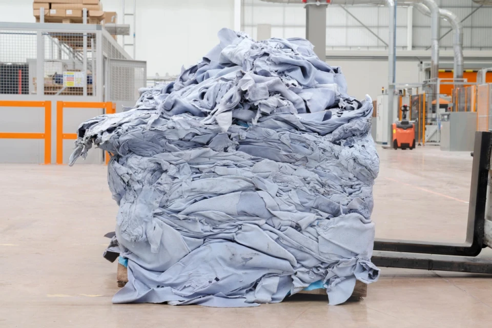 Large pile of folded light blue material on a wooden pallet in a factory or warehouse setting with a forklift nearby.