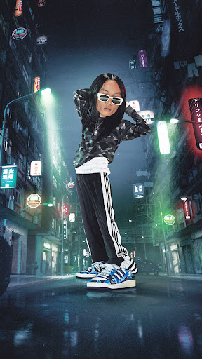 A person stands on a wet city street at night wearing a patterned hoodie, track pants, sneakers, and white sunglasses, with neon lights in the background.