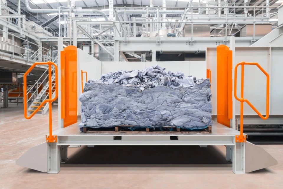 Industrial shot of a large bale of gray material on a loading platform, with machinery and metal structures in the background.
