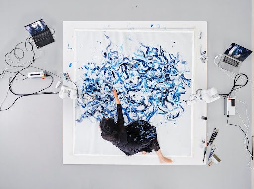 Overhead shot: woman in black skirt paints abstract blue swirls on a square canvas surrounded by robotic arms and laptops in a studio.