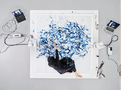 Overhead shot: woman in black skirt paints abstract blue swirls on a square canvas surrounded by robotic arms and laptops in a studio.