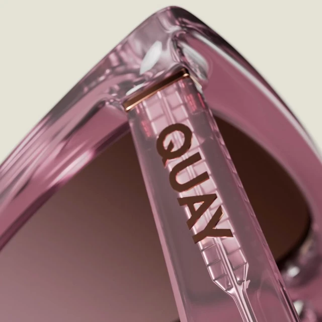 Closeup of pink Quay sunglasses arm displaying the logo. The sunglasses exhibit a translucent texture against a neutral background.