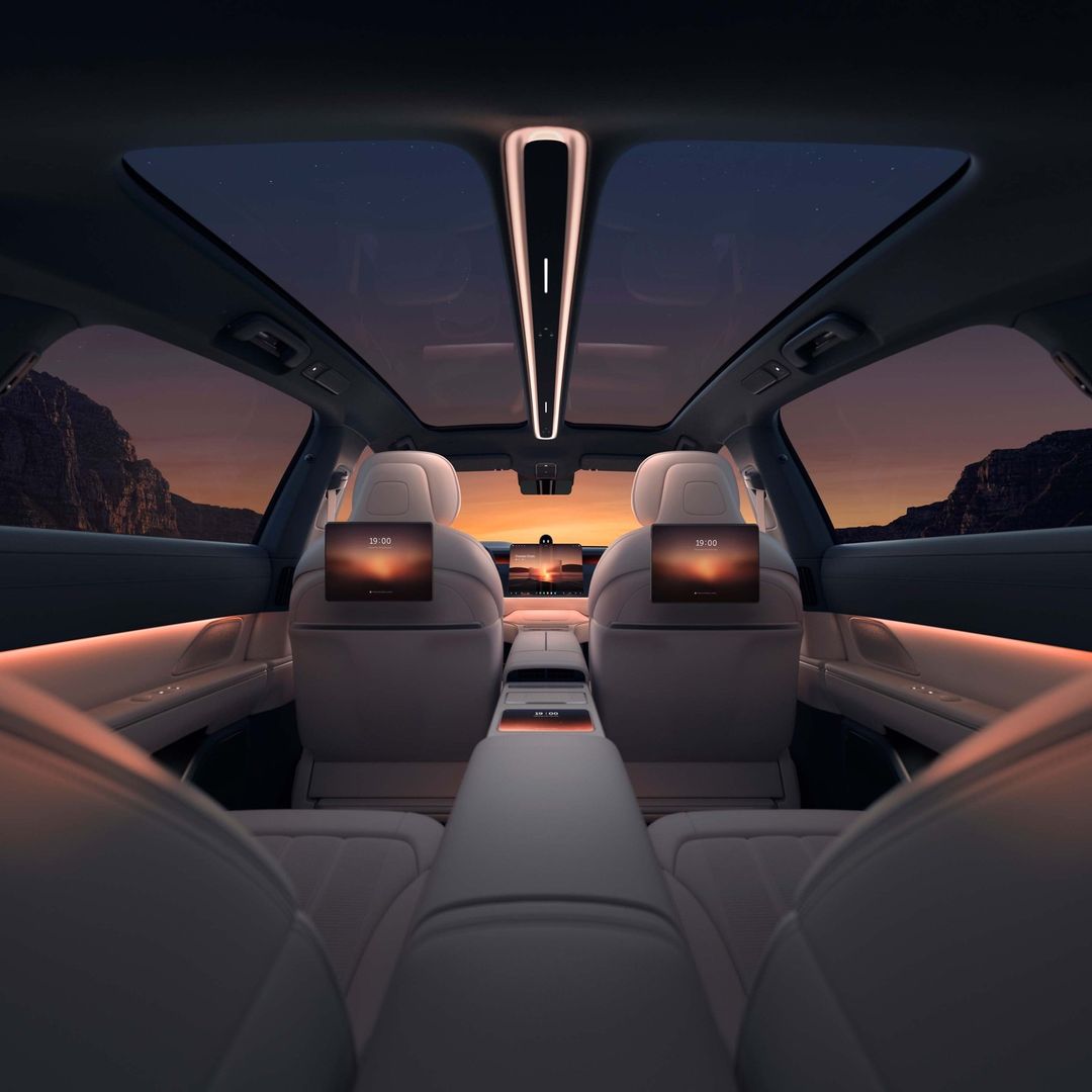 Car interior view with screens on back of seats showing sunset; the moon is visible through the sunroof and back window at dusk.