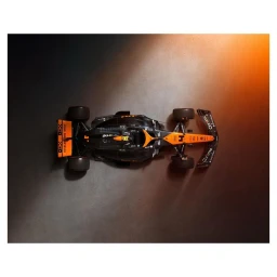 Top-down overhead view of a black and orange Formula 1 racing car on a textured dark studio floor with warm directional lighting.