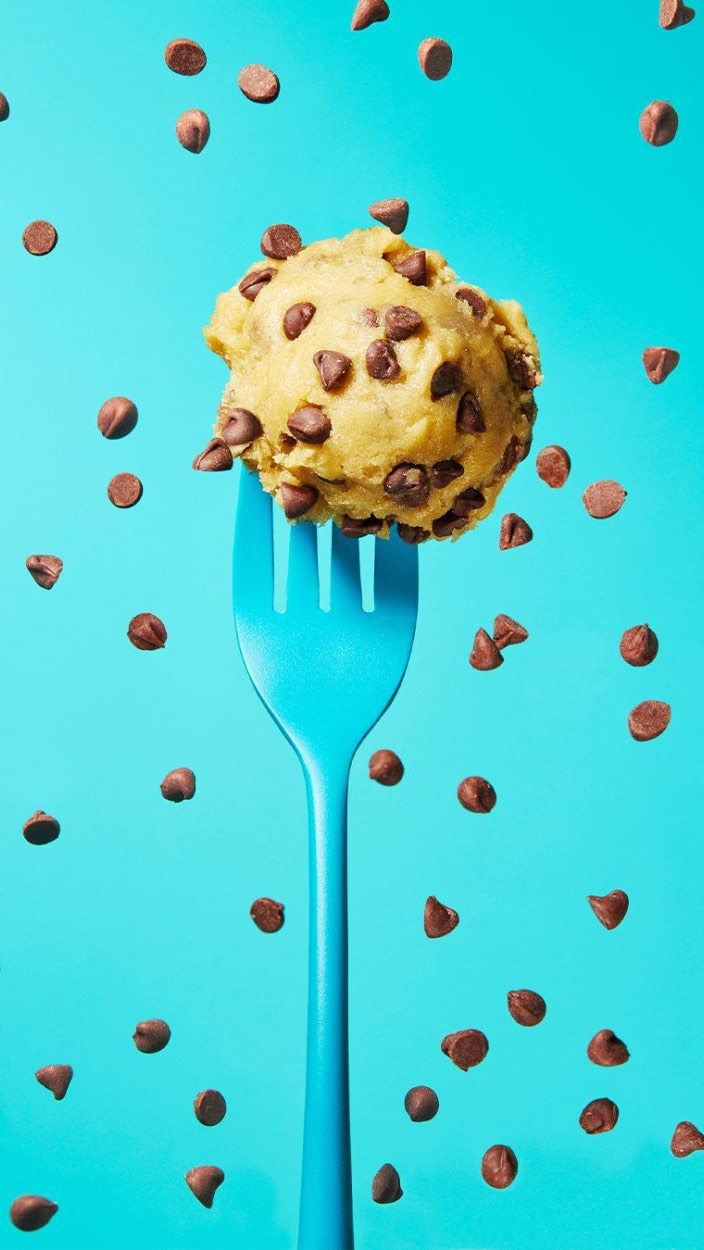Overhead shot of cookie dough ball topped with chocolate chips on blue plastic fork with chocolate chips scattered on light blue background.
