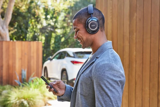 Man with headphones uses mobile phone outdoors near a modern car and wood fence; gray jacket, smiling; audio and mobile technology concept.