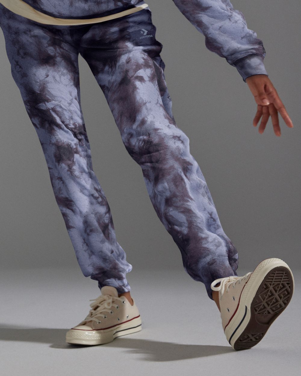 Person wearing tie-dye sweatpants and beige sneakers in a studio setting. The person's leg is bent and angled to the left.