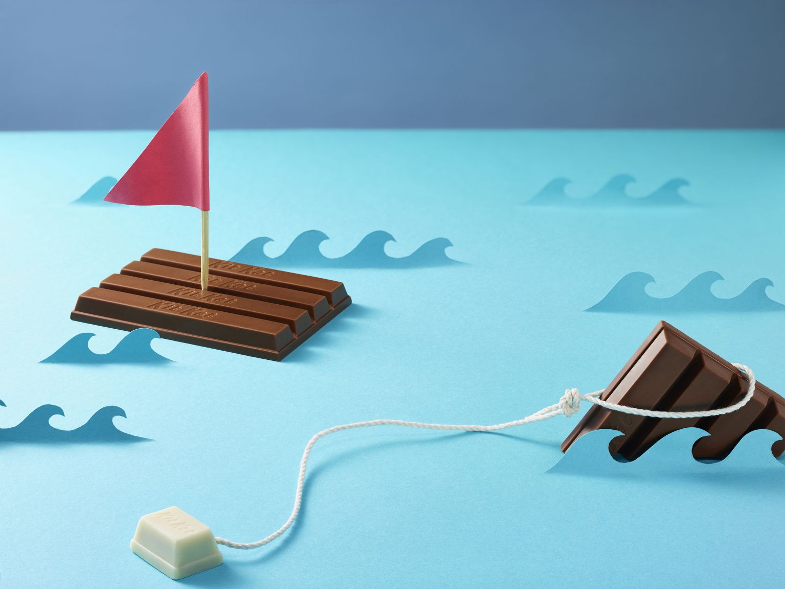 Creative still life of a Kit Kat chocolate bar crafted into boats on a blue paper sea with paper waves and a red paper sail.
