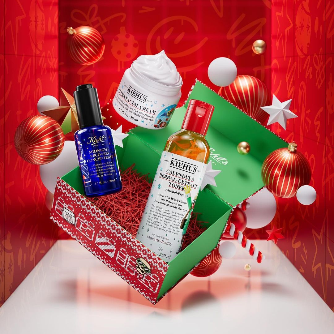 Kiehl's skincare products are displayed in a green gift box with red accents and confetti filling, against a red Christmas background.