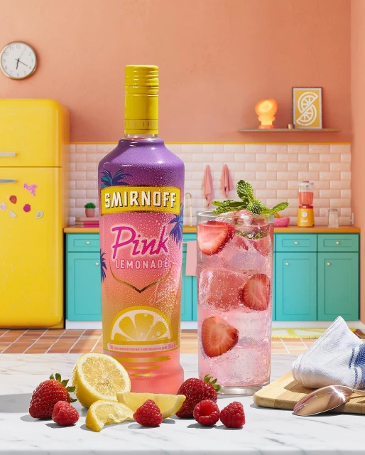 Still life shot of Smirnoff Pink Lemonade bottle next to a glass filled with the drink, ice, strawberries, mint, lemon and raspberries.
