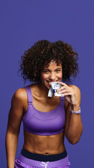 A smiling, athletic-build woman in workout attire with curly hair eats a snack bar against a solid purple backdrop in a medium shot.