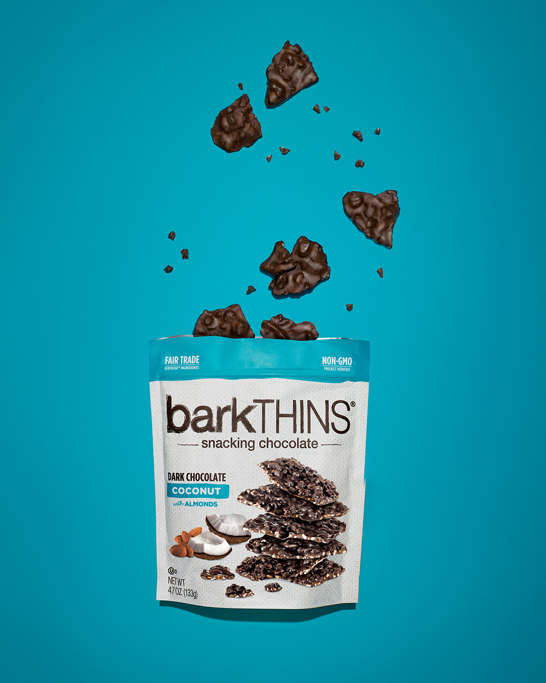 BarkTHINS snacking chocolate packaging with pieces of chocolate falling on top against a teal background, studio shot.
