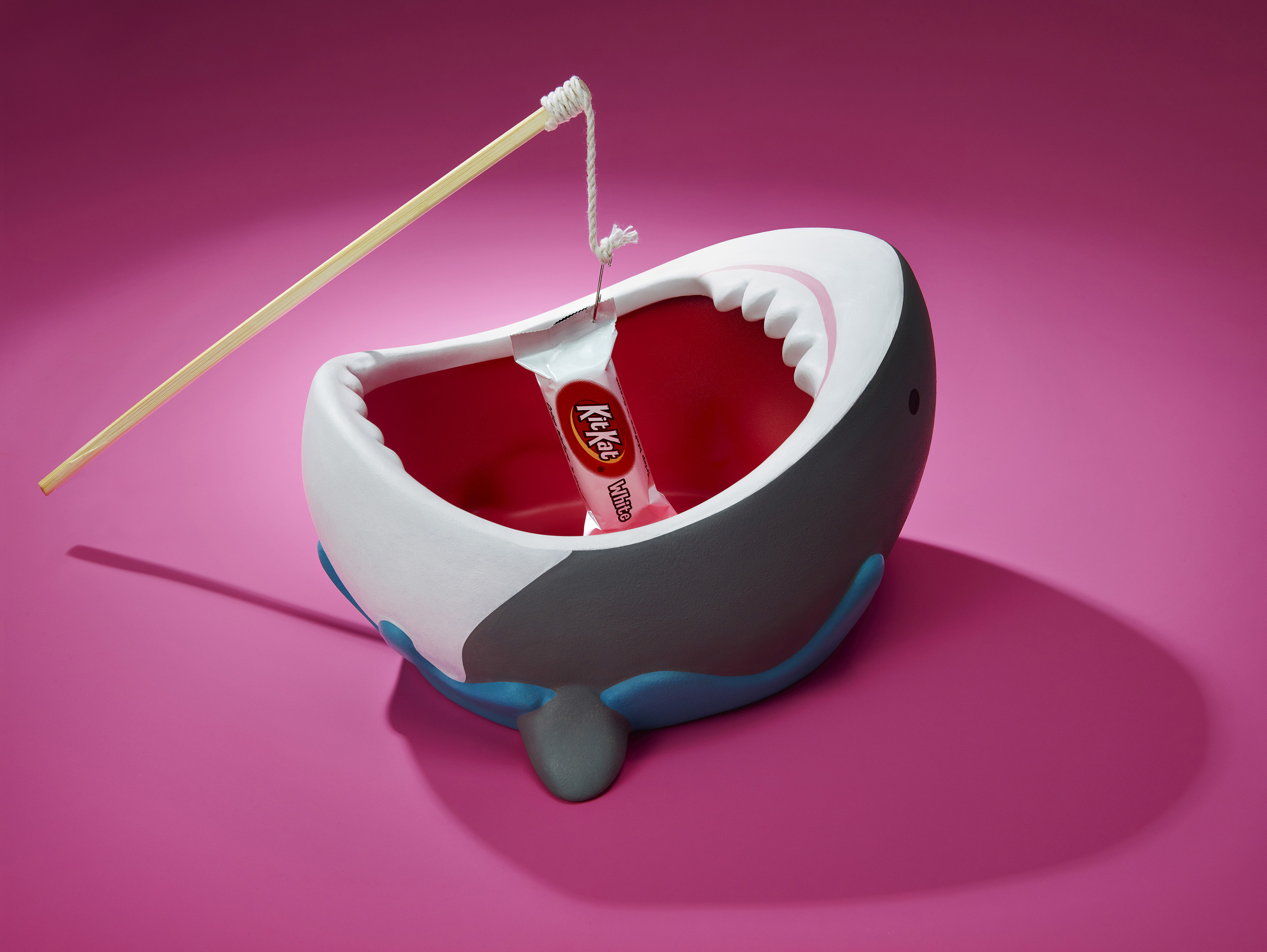 Whimsical studio shot of a KitKat candy bar 'caught' in a shark-shaped bowl with a fishing pole on a vibrant pink backdrop.