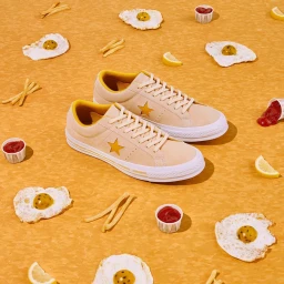 A pair of beige and yellow sneakers surrounded by fried eggs, french fries, and ketchup on a textured yellow background.