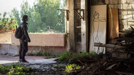 Person wearing dark jacket and backpack standing inside a ruined building looking at a whimsical line drawing on a wooden panel.