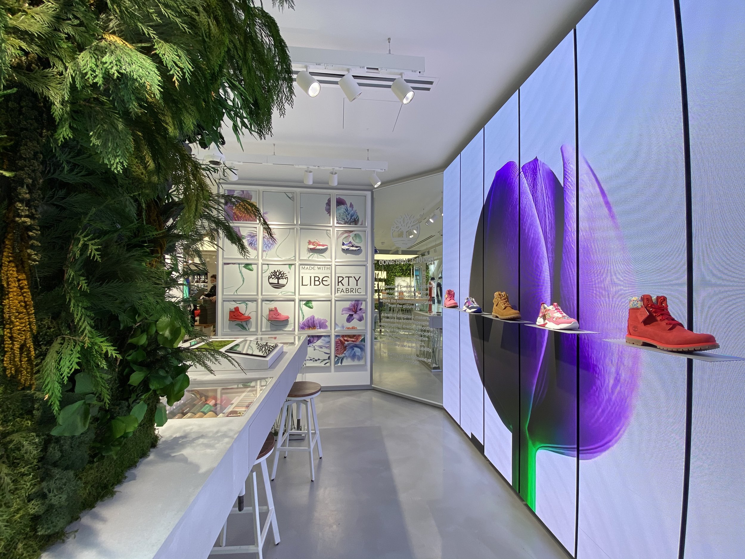 Interior of a shoe store with a vertical garden, white display fixtures, and an electronic display showing a purple flower.