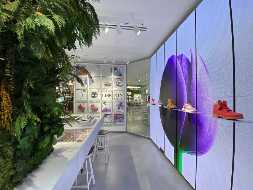 Interior of a shoe store with a vertical garden, white display fixtures, and an electronic display showing a purple flower.